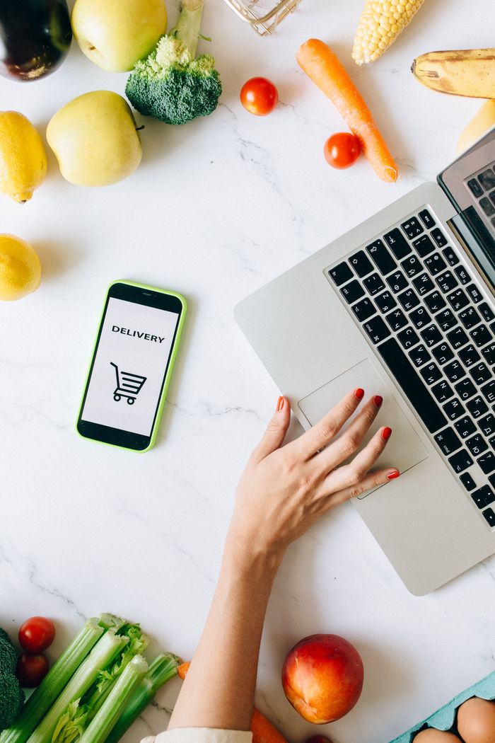 Services A top view of online grocery shopping using a laptop and smartphone surrounded by fresh vegetables and fruits.